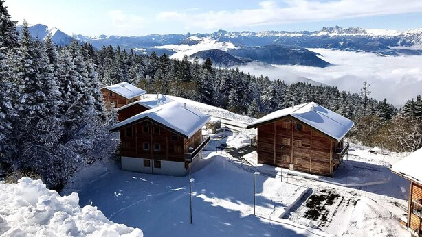 Un chalet de montagne enneigé, doté de plusieurs cabanes en bois, entouré de forêts denses et d’arbres recouverts de neige, et offrant une vue panoramique sur les montagnes et les vallées lointaines.