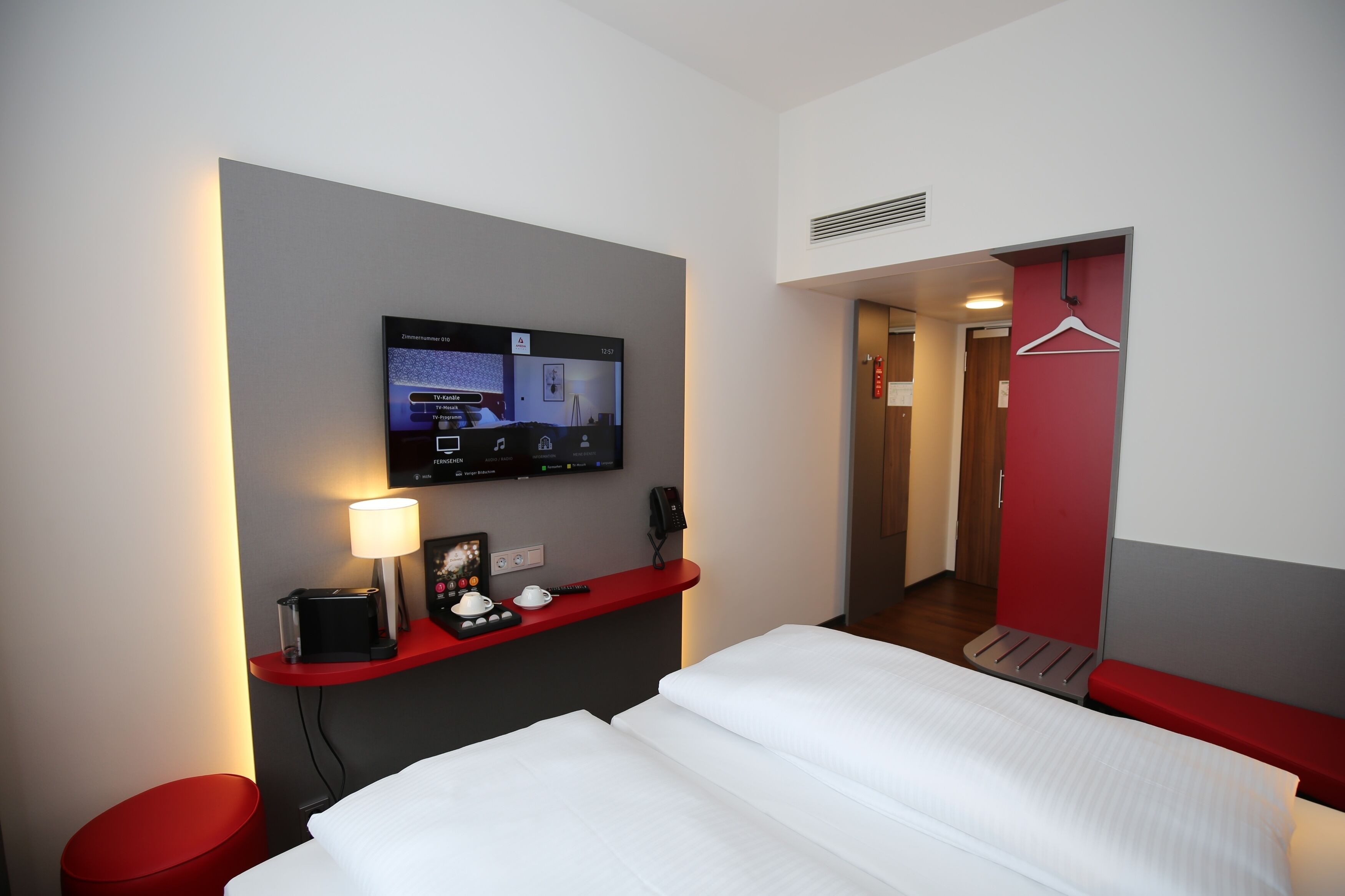 Hotellihuone, jossa on sänky, seinälle asennettu televisio, punainen penkki, pieni pöytä lampun kanssa ja punainen seinä.