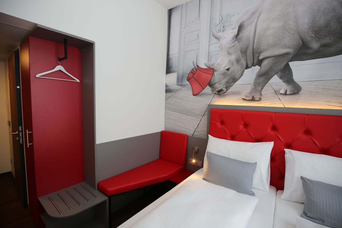 Habitación de hotel con una decoración en tonos rojo y gris, una cama con ropa de cama blanca y gris, un banco tapizado en rojo y un mural en la pared con un rinoceronte.