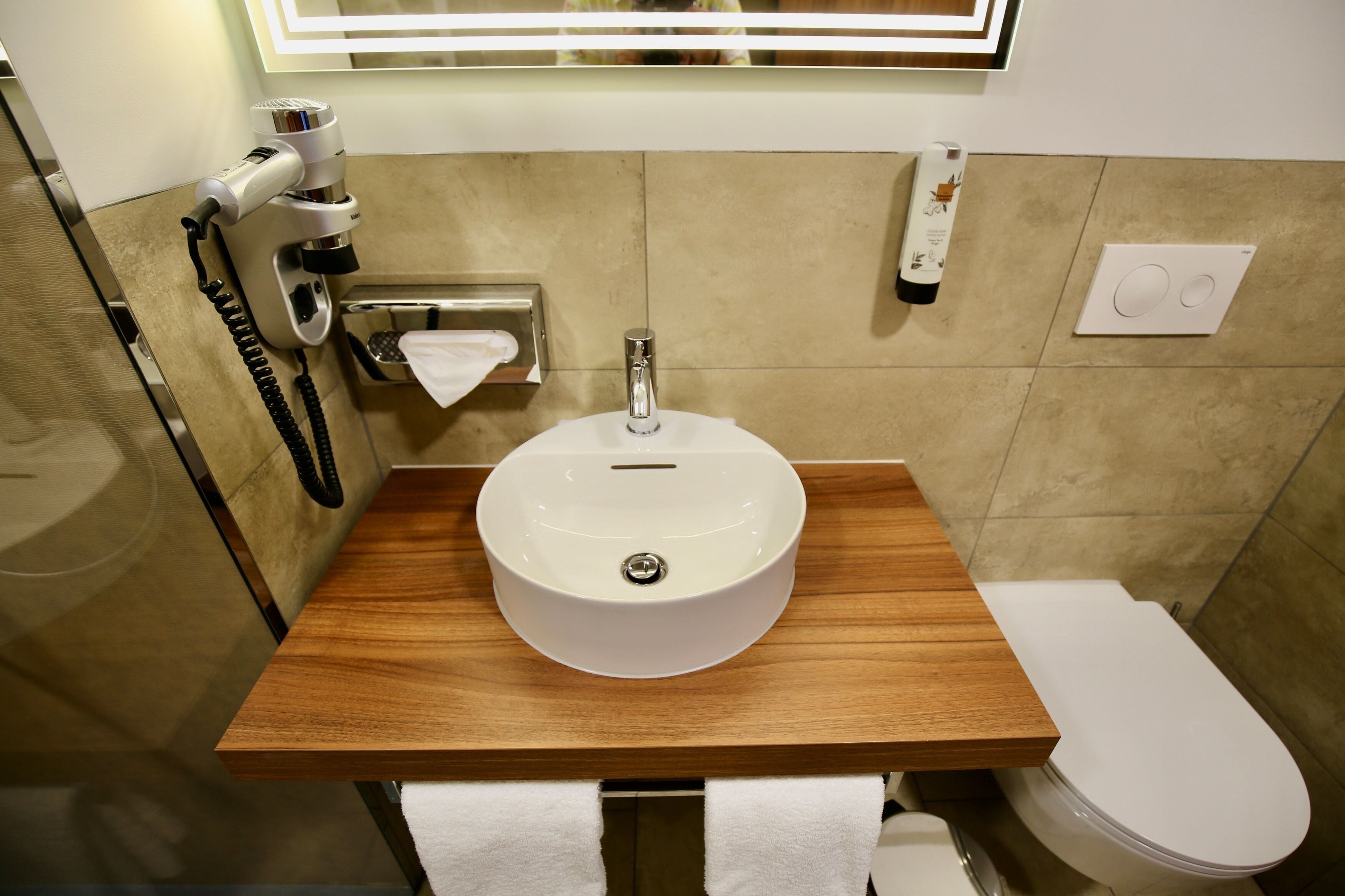 A bathroom with a wooden vanity, a white sink, a toilet, and a hairdryer on the wall.