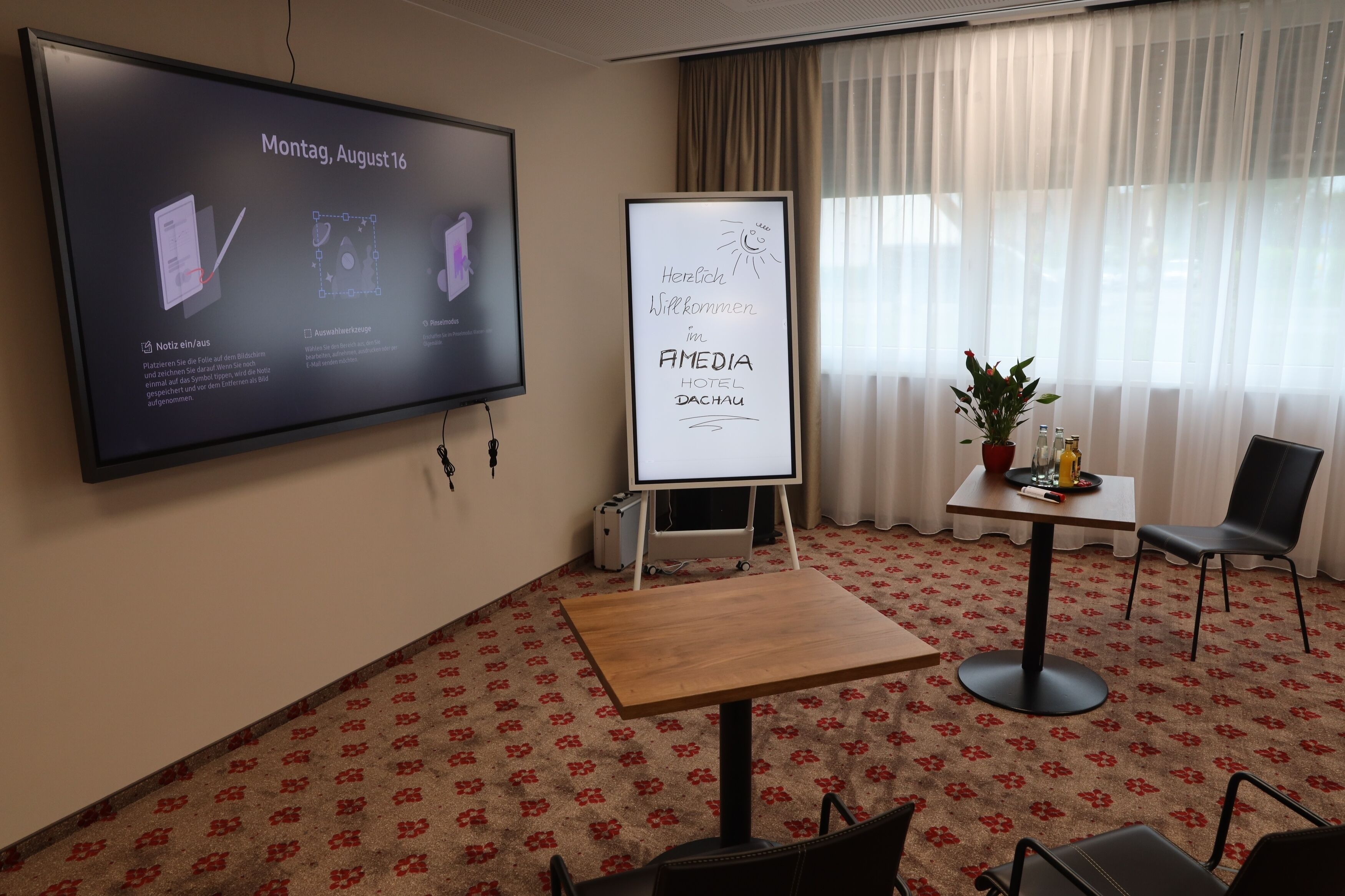 A hotel room with a large screen displaying a presentation, a whiteboard with a welcome message, a small table with a plant, and a chair.