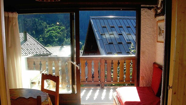 Et værelse med en træstol, et lille bord, en rød seng og en balkon med udsigt til huse og træer.