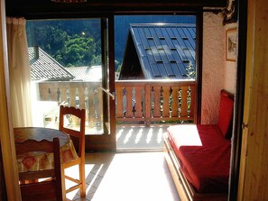 A room with a wooden chair, a small table, a red bed, and a balcony with a view of houses and trees. - Champagny-en-Vanoise: Studio 4 ppl, balcony, pets allowed (Champagny-en-Vanoise)