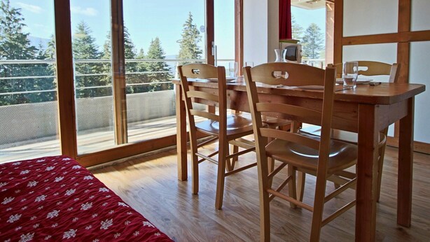 Ein Zimmer mit einem Holzespeisetisch und Stühlen, einem Bett mit rotem, gemustertem Überwurf und großen Fenstern mit Blick auf Bäume und einen Balkon.