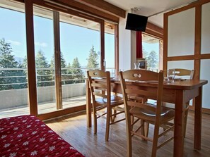 A room with a wooden dining table and chairs, a bed with a red patterned bedspread, and large windows offering a view of trees and a balcony.