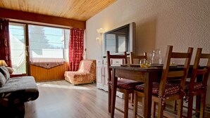 Une pièce chaleureuse avec un plafond en bois, une table à manger avec des chaises, un canapé et un fauteuil agrémenté d’un coussin à motifs.