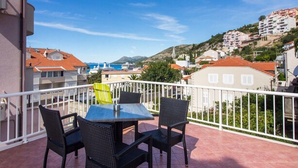 Un balcon avec une table et des chaises, donnant sur une ville côtière avec ses bâtiments et son église.