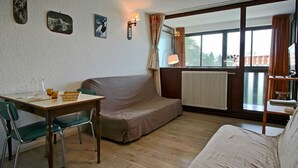 A room with a wooden floor, a sofa, a table with chairs, a radiator, and a window with curtains. - Bright studio with loggia near slopes and shops (Chamrousse)