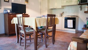 Een eetgedeelte met houten stoelen en een tafel, een televisie op een kast, en een keuken met witte kastjes en een fornuis.