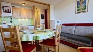 A dining area with a table set for four, surrounded by wooden chairs with red cushions. A sofa is visible in the background. - Bright apartment near slopes, 2 rooms, West-facing balcony (Chamrousse)