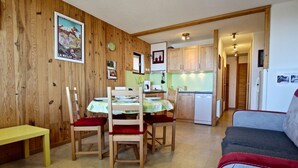 A cozy living room with a dining table, chairs, and a sofa. A kitchen with wooden cabinets and a refrigerator is visible in the background. - Bright apartment near slopes, 2 rooms, West-facing balcony (Chamrousse)