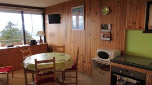 A cozy cabin interior with a wooden interior, a dining table with chairs, a kitchen area with a microwave and oven, and a television mounted on the wall. - Bright apartment near slopes, 2 rooms, West-facing balcony (Chamrousse)