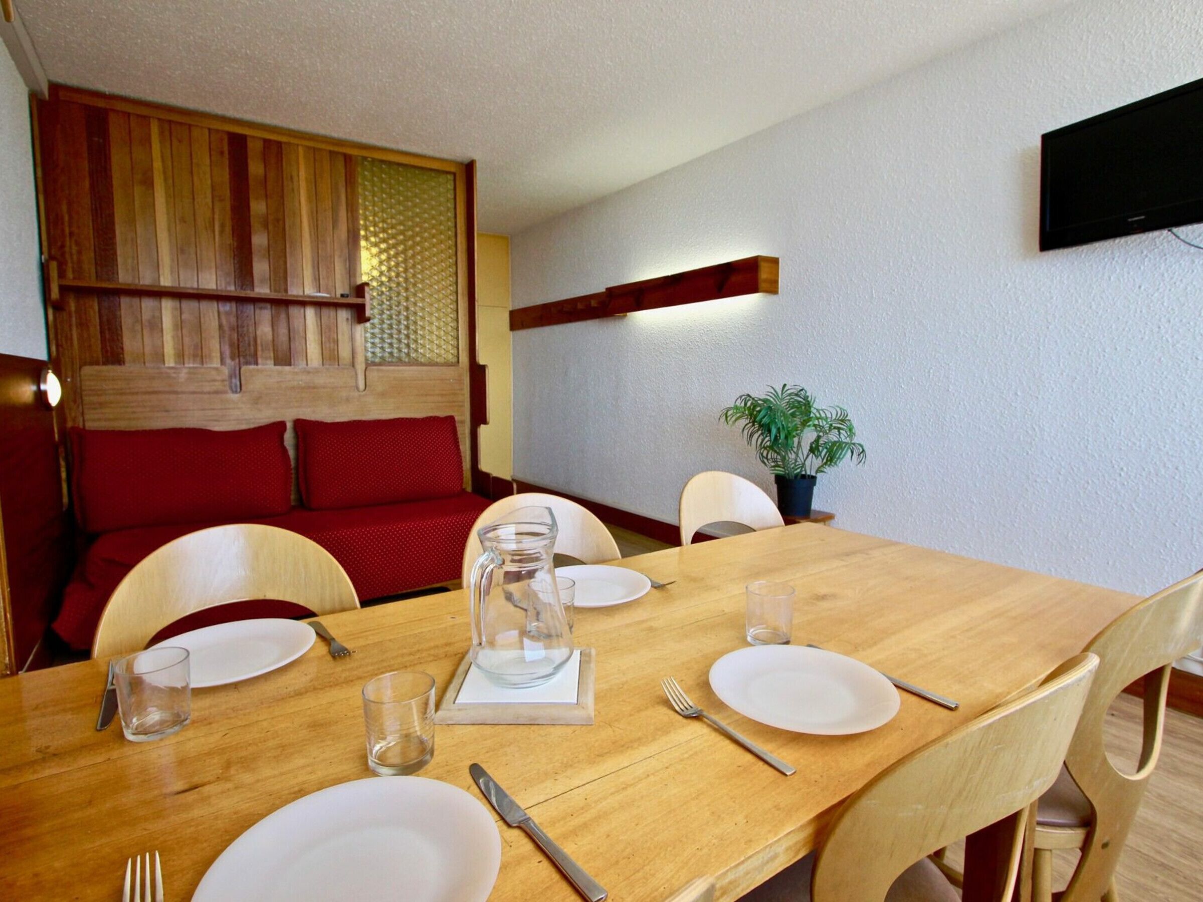 A dining area with a wooden table set for four, surrounded by chairs. There is a television mounted on the wall, a potted plant on a small table, and a red sofa in the background.