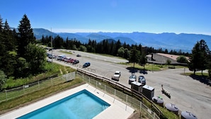 Ein Schwimmbad mit Bergblick, ein Parkplatz und ein Gebäude mit einer gewölbten Dachform.