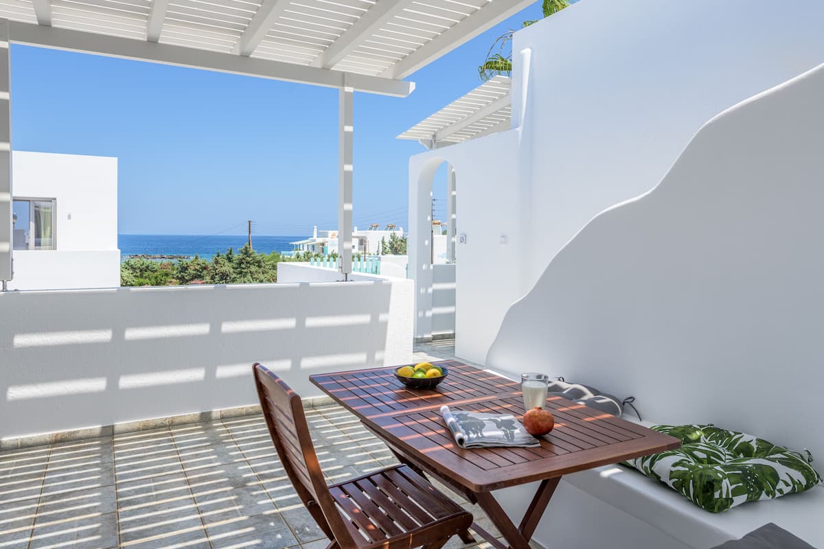 A wooden table with a bowl of fruit, a glass of milk, and a newspaper on a balcony with a view of the sea.