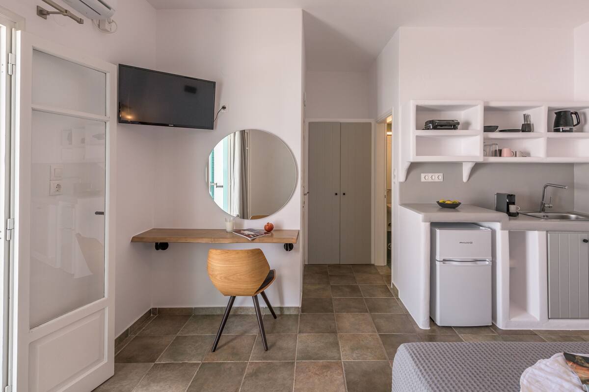 A modern hotel room with a wooden desk, a small chair, a flat-screen TV, a compact kitchenette, and a wardrobe.
