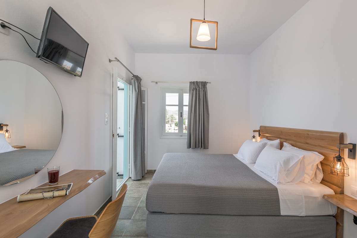 A modern hotel room with a large bed, a bedside table with a lamp, a mirror, and a view of the outdoors through a window.