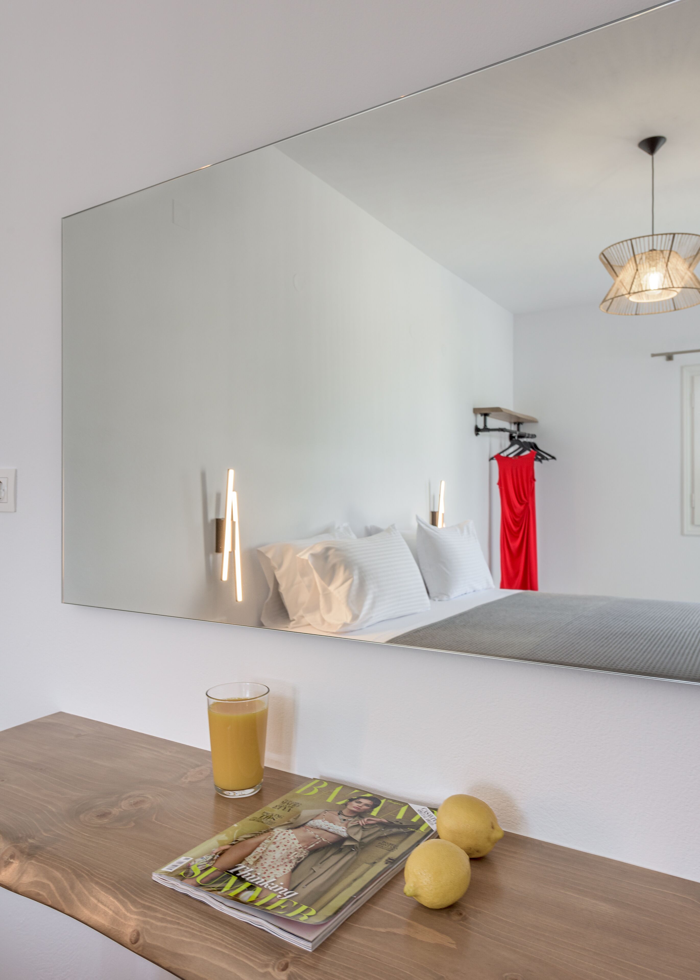 Una habitación de hotel con un espejo grande, una mesa de madera con una revista y limones, un vaso de zumo de naranja y una cortina roja.