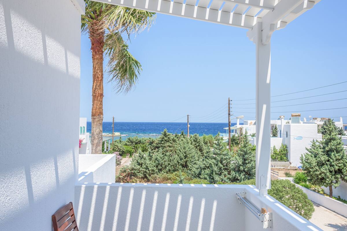 A balcony with a wooden table and chairs, a white pergola, and a view of the sea and greenery.