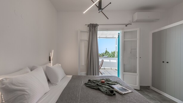 A modern bedroom with a bed, a bedside table, a window with curtains, and an open door leading to a balcony. - Michalis Studios (Milos)