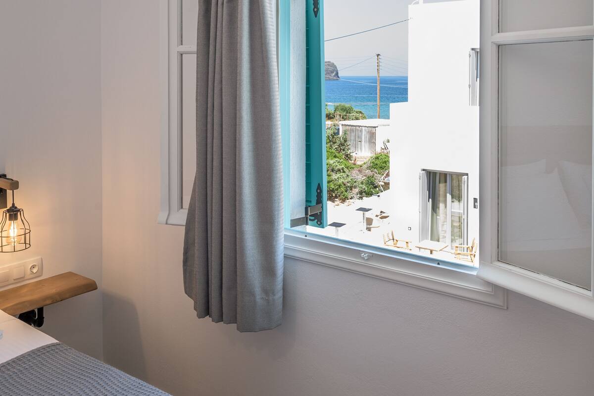 A room with a window overlooking a coastal view, a bed, a bedside table, and a lamp.