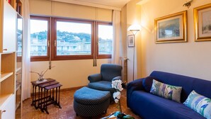 Un soggiorno con un divano blu, un poggiapiedi decorato, un tavolino con un vaso e vista sugli edifici attraverso ampie finestre.