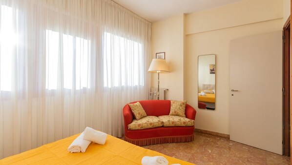 A hotel room with a bed, a red sofa, a lamp, a mirror, and a door.