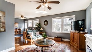 A spacious living room with a ceiling fan, a television, a fireplace, and a large window. - Dream Catcher Bungalow - Near Uptown, Bojangles Coliseum & BOA! Pet & Family Friendly, Plus 15% off! (Charlotte)