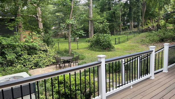 Eine Terrasse mit weißer Geländer, die auf einen Garten mit Rasen, Bäumen und einer Terrasse mit Gartenmöbeln blickt.