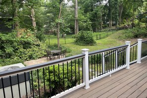 A deck with a white railing overlooking a backyard with a lawn, trees, and a patio with outdoor furniture.