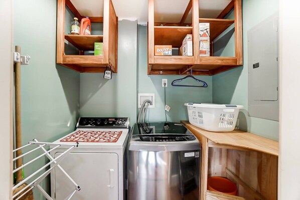 A compact kitchen with a stove, washing machine, and drying rack. There are wooden shelves above holding various items.