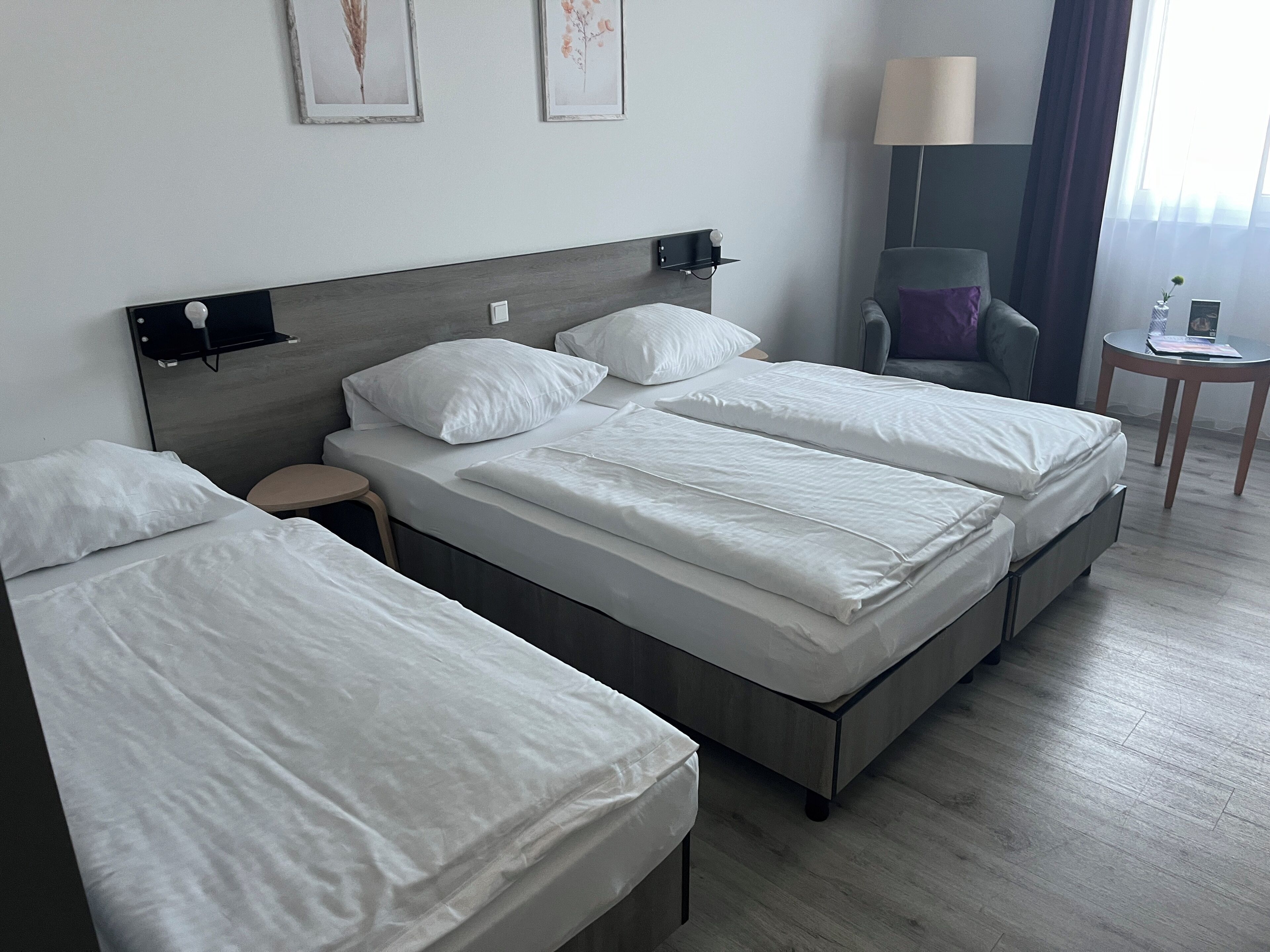 A hotel room with a double bed, a single bed, a small table, a chair, and a lamp.