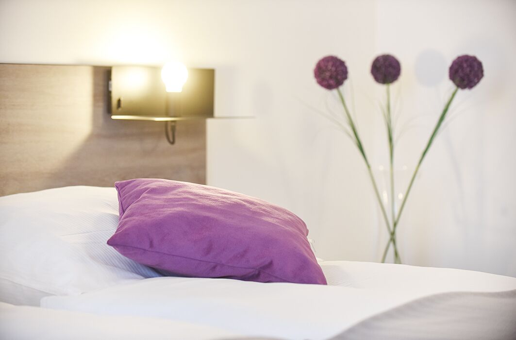Ein Bett mit einem violetten Kopfkissen und einer Wandleuchte.
