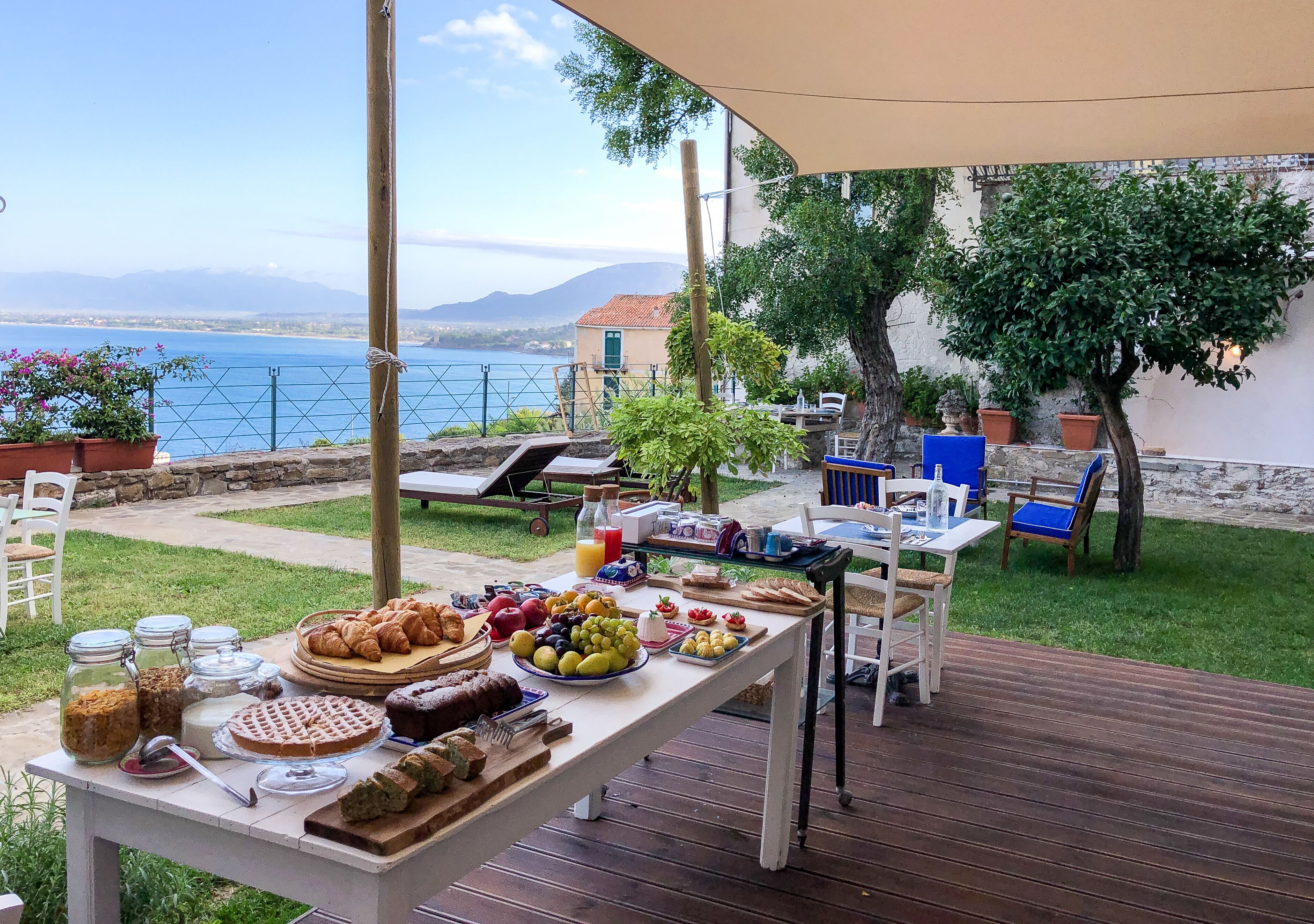 Un desayuno al aire libre con una mesa repleta de pasteles, frutas y bebidas, con vistas a un lago y montañas panorámicas.