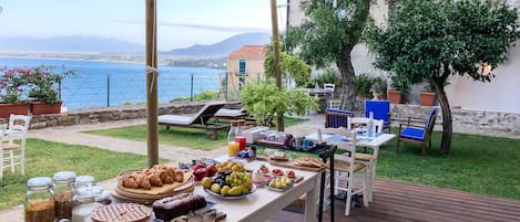 Un desayuno al aire libre con una mesa repleta de pasteles, frutas y bebidas, con vistas a un lago y montañas panorámicas.