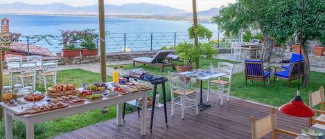 An outdoor dining area with a view of the sea and mountains, wooden deck flooring, and various dining tables and chairs.