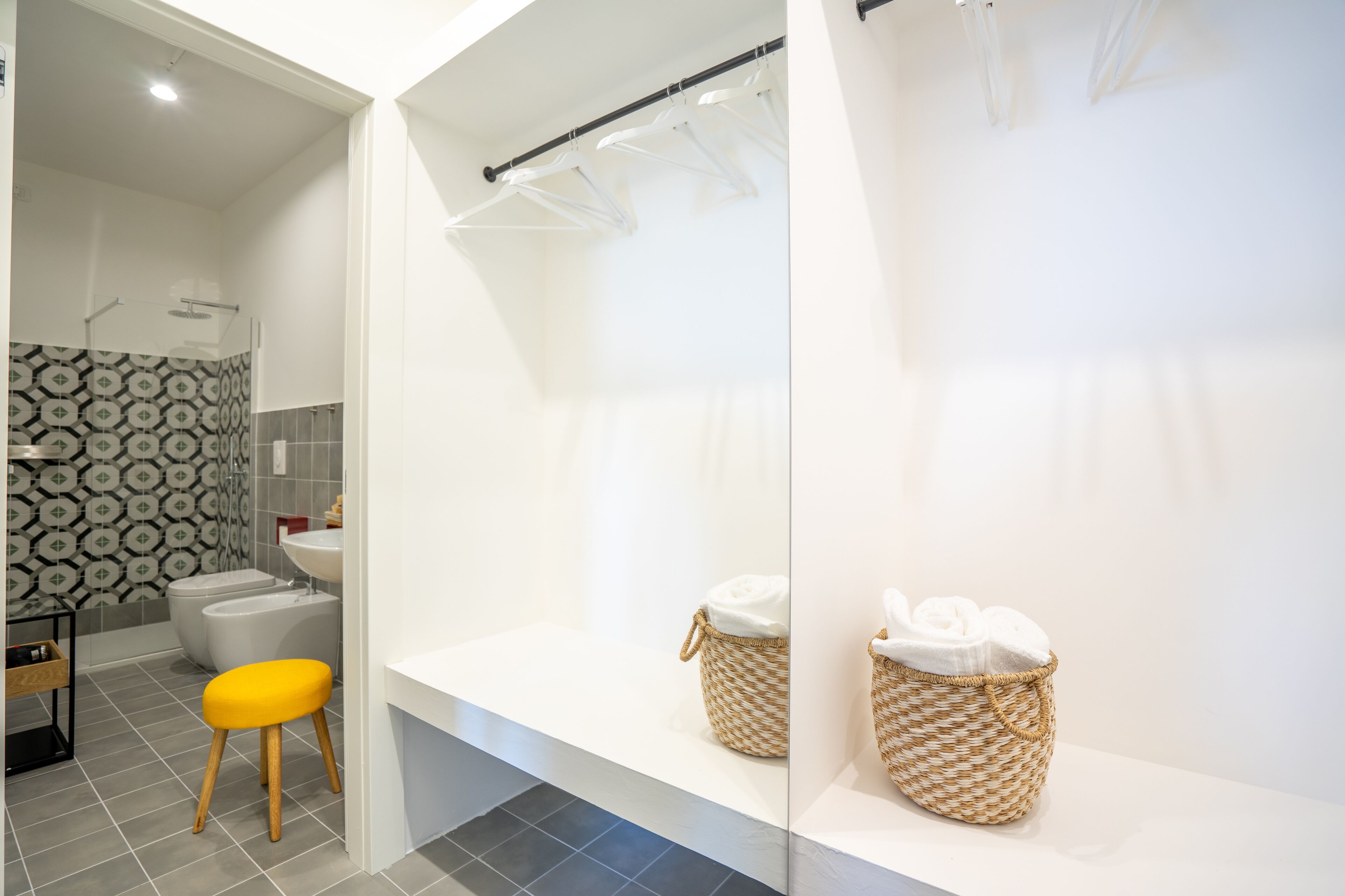 A modern bathroom with a white sink, two wicker baskets on a white shelf, a yellow stool, and a patterned wall.