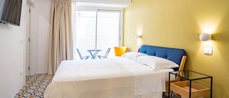 Un dormitorio con cama, una mesita de noche, una silla, una ventana con cortinas y un televisor montado en la pared.