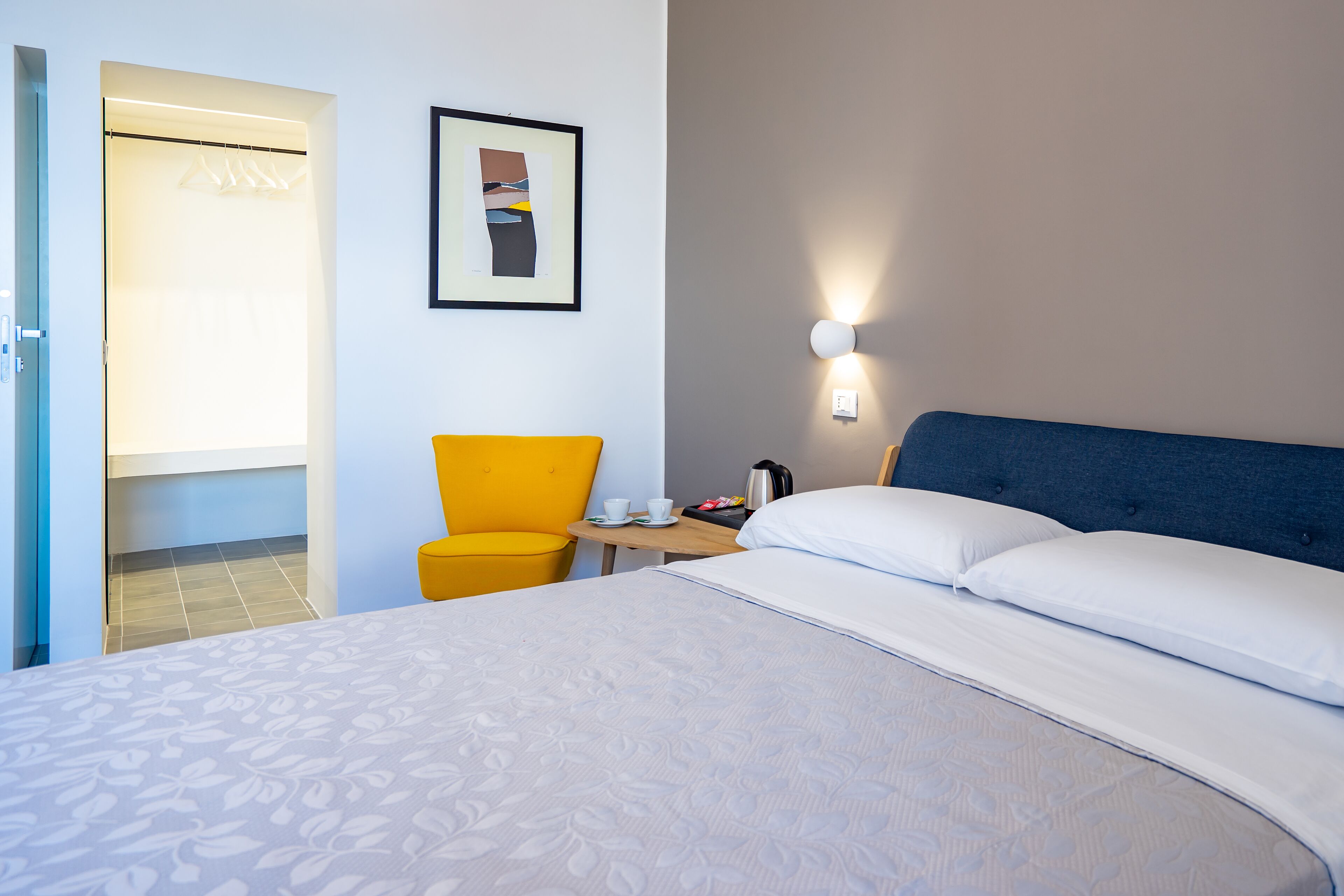 Habitación de hotel con una cama grande, una mesita de noche con una taza y una tetera, una silla y un cuadro en la pared.