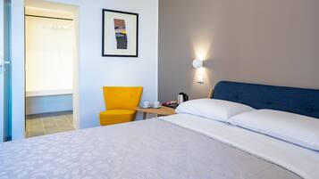Habitación de hotel con una cama grande, una mesita de noche con una taza y una tetera, una silla y un cuadro en la pared.