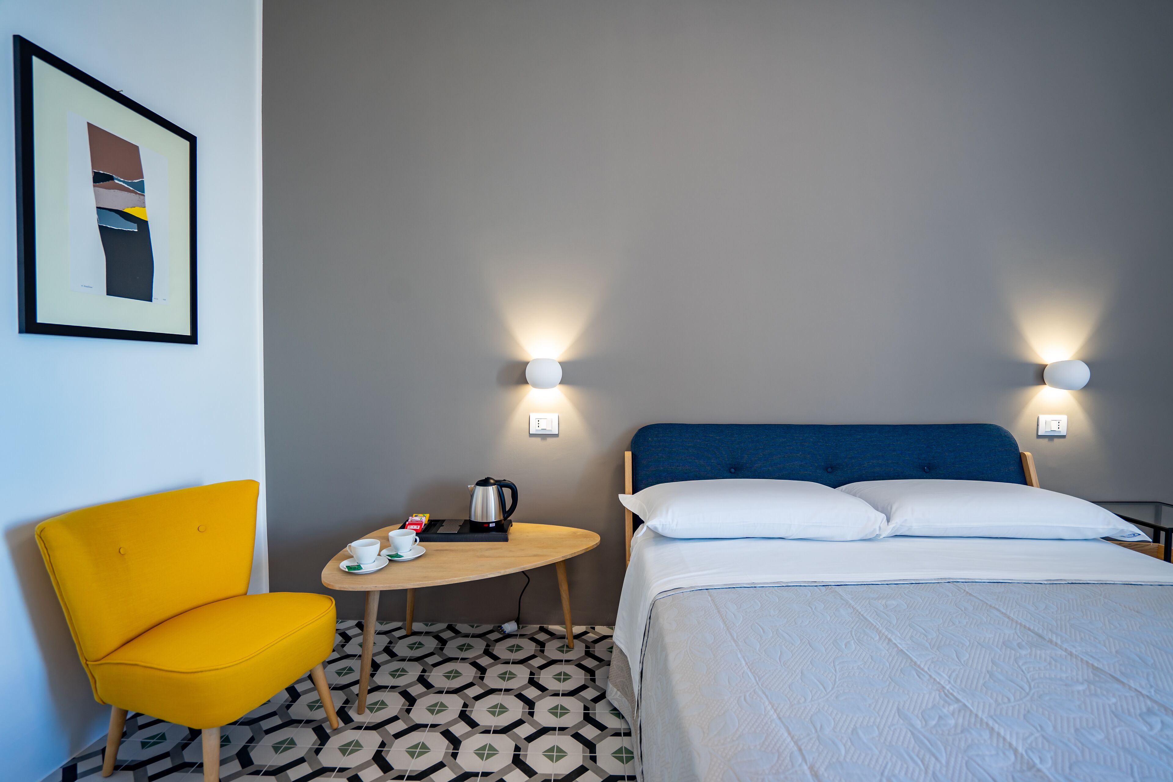 A hotel room with a bed, a yellow chair, a small table with a kettle, and two wall-mounted lights.