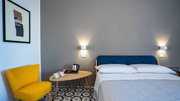 Une chambre d’hôtel avec un lit, une chaise jaune, une petite table avec une bouilloire et deux lampes fixées au mur.