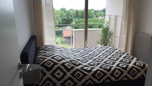 Un dormitorio con una cama cubierta por una colcha estampada, una ventana que ofrece vistas a zonas verdes y edificios, y un radiador en la pared.