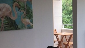 Un comedor con mesa de madera y sillas negras, un cuadro de flamencos en la pared y vistas a la vegetación a través de un vano abierto.