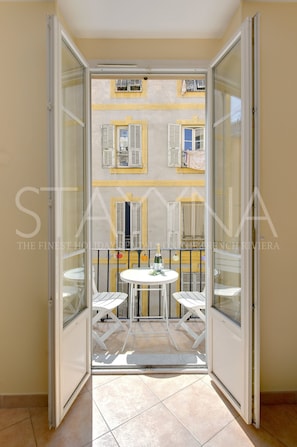 A balcony with a table and chairs, a view of a building with shutters, and a tiled floor. - Juliette 1 bedroom apartment VRBO (Nice)