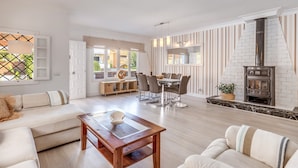 Een ruime woonkamer met een bank, eettafel en stoelen, een open haard en uitzicht door grote ramen.
