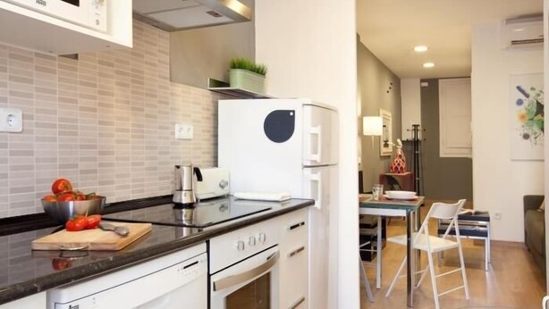 A modern kitchen with white appliances, a glass dining table, and a view into the living area. - Sagrada Familia - Apartment in Barcelona (Barcelona)