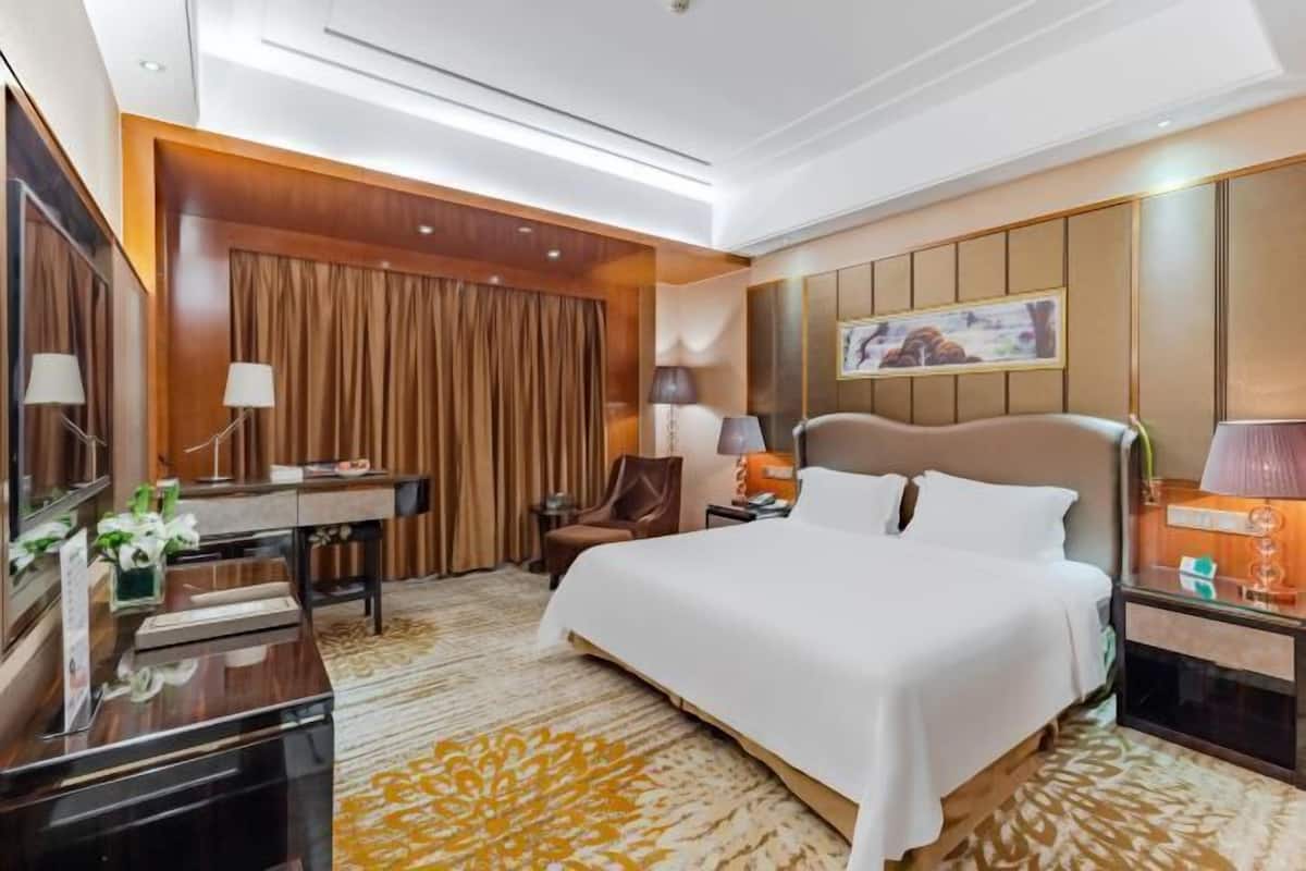 A hotel room with a large bed, a desk, a chair, and a nightstand with a lamp.