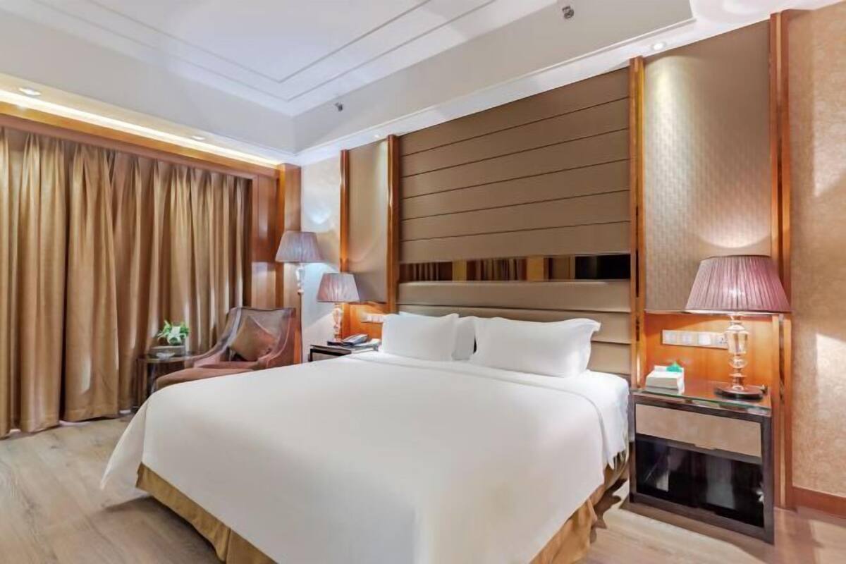A hotel room with a large bed, wooden headboard, two bedside lamps, a nightstand, and a curtain.