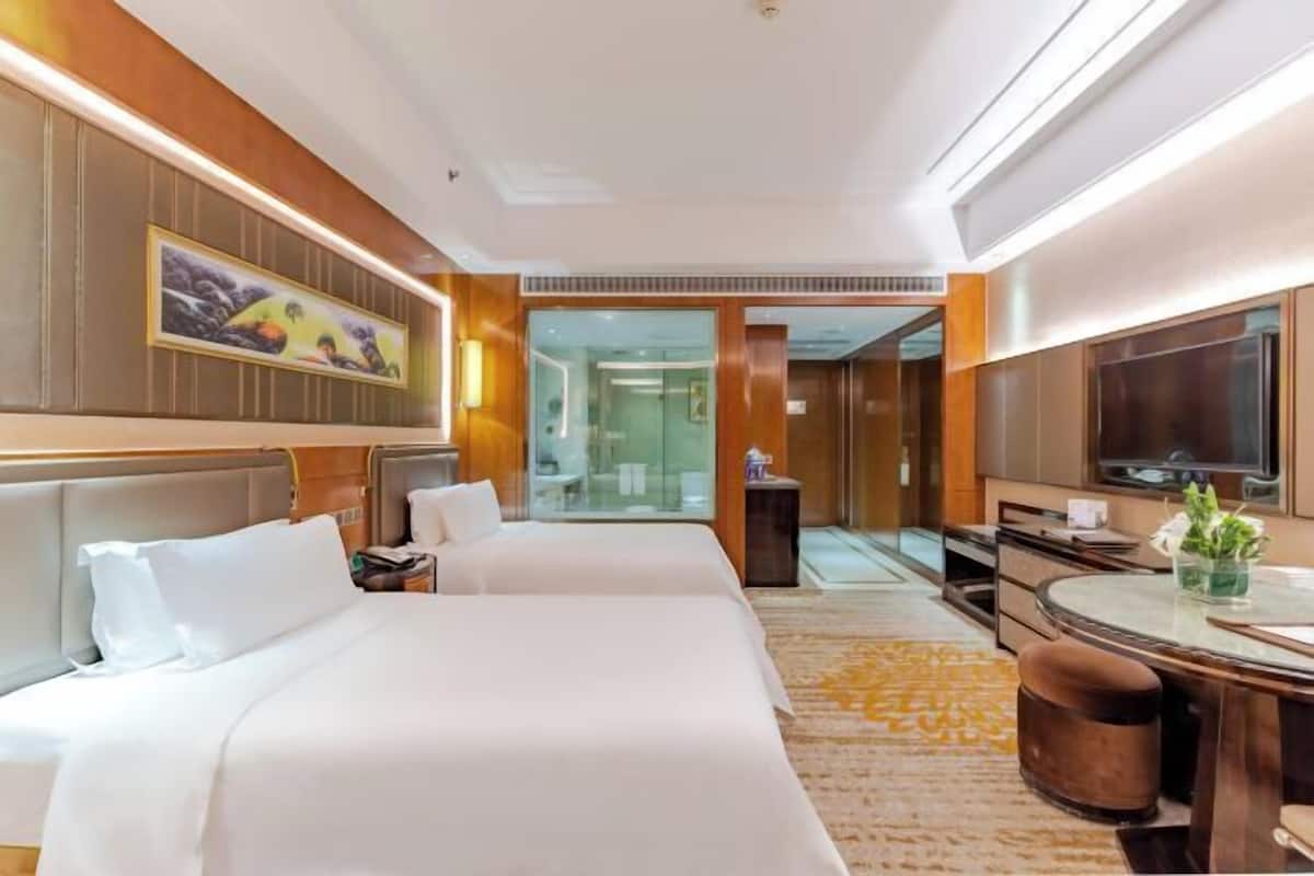 A hotel room with two beds, a desk, a television, and a bathroom visible through a glass door.